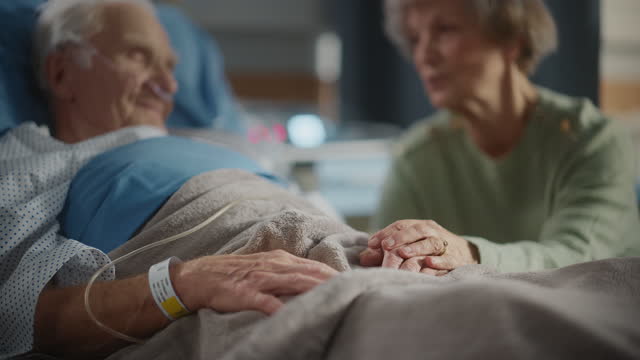 Hospital Ward: Focus on Hands of Elderly Man Resting in Bed, His Caring Wife Supports Him By Sitting Beside and Comforting with Her Touch. Old Man Recovering successfuly after Sickness and Surgery